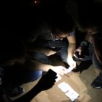 Trzy osoby w ciemności z pomocą latarek probują odczytać tekst z kartek papieru.