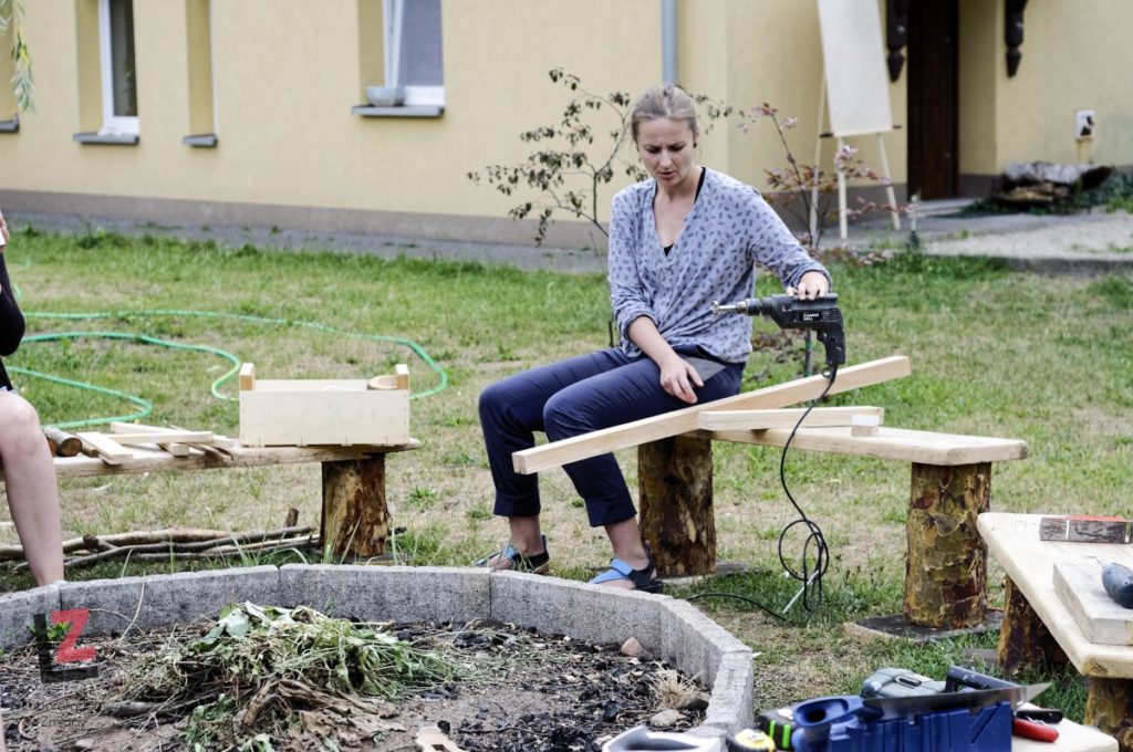 kobieta siedząca na ławce przy kręgu do ogniska z pomocą wiertarki zespala drewniane elementy.